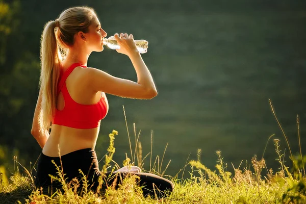 Una giovane bionda con un top rosso e pantaloni neri si siede sull'erba in natura. Una donna sportiva tiene in mano una bottiglia d'acqua . — Foto Stock