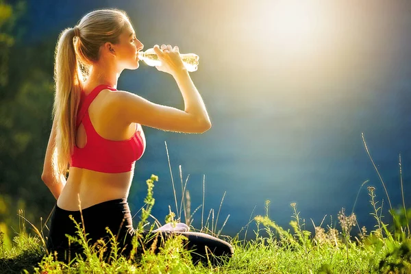 Una giovane bionda con un top rosso e pantaloni neri si siede sull'erba in natura. Una donna sportiva tiene in mano una bottiglia d'acqua . — Foto Stock
