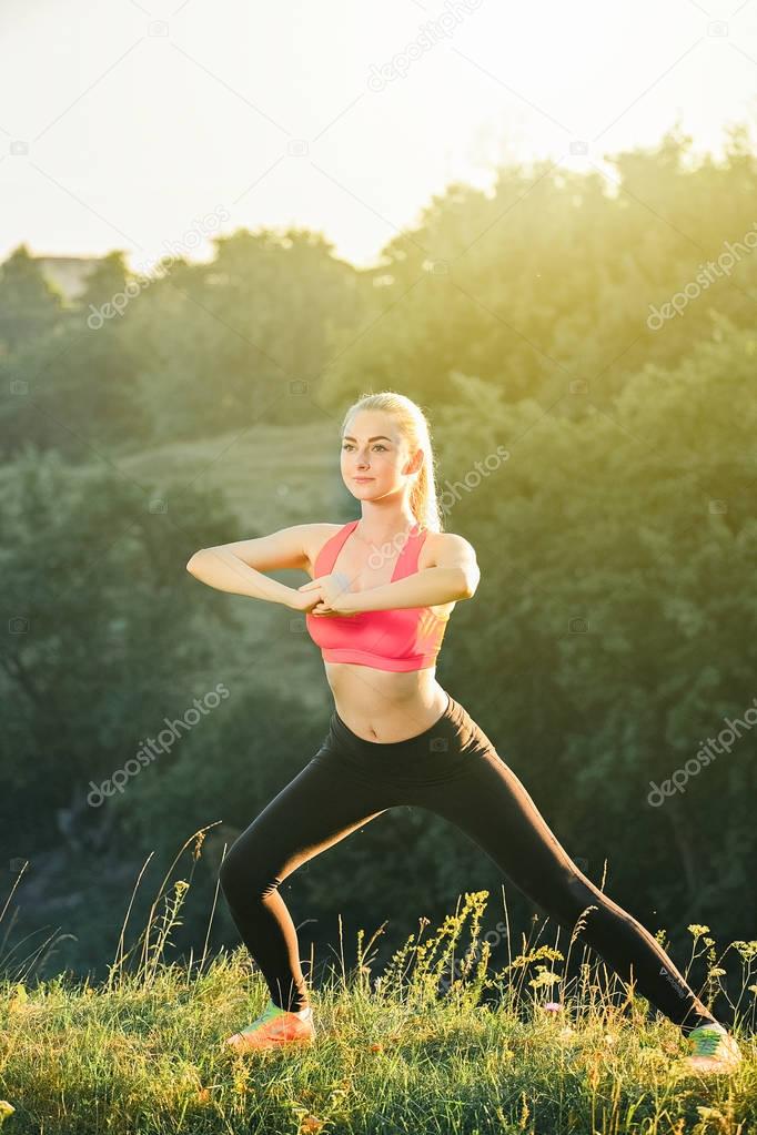 A young blonde in a red top and black pants doing sports exercises in nature.