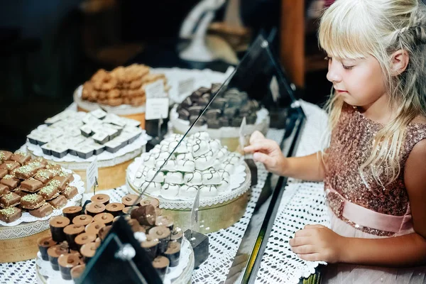 A little blonde girl is standing near a shop window with an assortment of sweets. The child chooses sweets