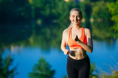doğa üzerinde çalışan genç sarışın bir kadın. Bir kırmızı bir spor kız T-shirt Nehri yakınında park boyunca çalışır.