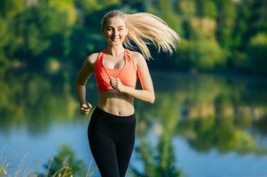 doğa üzerinde çalışan genç sarışın bir kadın. Bir kırmızı bir spor kız T-shirt Nehri yakınında park boyunca çalışır.