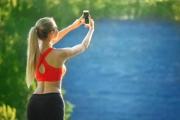 Młoda blond kobieta stoi na tle błękitnego morza. Kobiety sporty w kolorze czerwonym t-shirt sprawia, że selfie na natury, w pobliżu morza — Zdjęcie stockowe