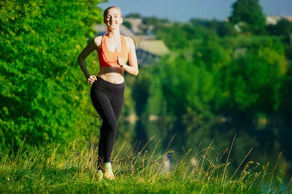 Joven rubia corriendo sobre la naturaleza. Una chica deportiva con una camiseta roja corre por el parque cerca del río . — Foto de Stock