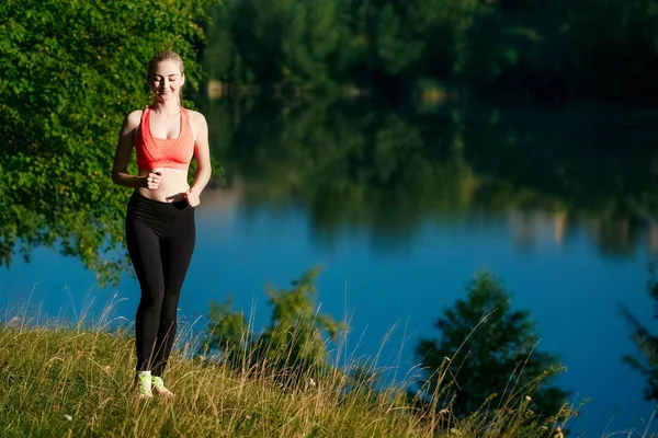doğa üzerinde çalışan genç sarışın bir kadın. Bir kırmızı bir spor kız T-shirt Nehri yakınında park boyunca çalışır.