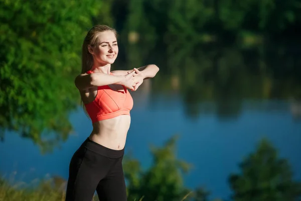 Joven rubia haciendo ejercicios en la naturaleza. Una chica deportiva en un chaleco rojo está de pie en un parque cerca del río — Foto de Stock