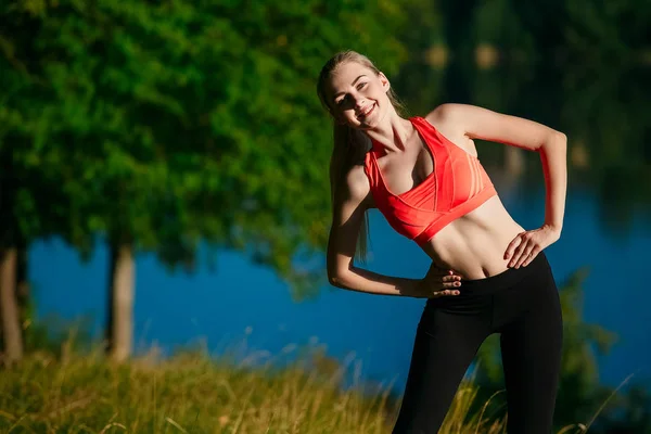 Giovane donna bionda che fa esercizi in natura. Una ragazza sportiva in gilet rosso è in piedi in un parco vicino al fiume — Foto Stock