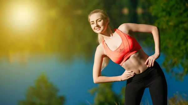Giovane donna bionda che fa esercizi in natura. Una ragazza sportiva in gilet rosso è in piedi in un parco vicino al fiume — Foto Stock