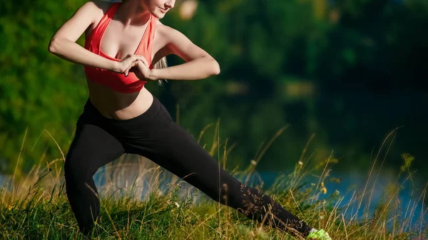 Young blond woman doing exercises in nature. A sports girl in a red vest is standing in a park near the river — Stock Photo, Image