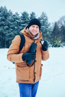 kar üzerinde duran ve bir termos ve bir fincan çay tutan bir sırt çantası ile kırmızı ceketli genç bir adam. Kış orman.