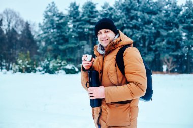 kar üzerinde duran ve bir termos ve bir fincan çay tutan bir sırt çantası ile kırmızı ceketli genç bir adam. Kış orman.