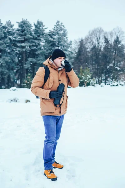 kar üzerinde duran ve bir termos ve bir fincan çay tutan bir sırt çantası ile kırmızı ceketli genç bir adam. Kış orman.