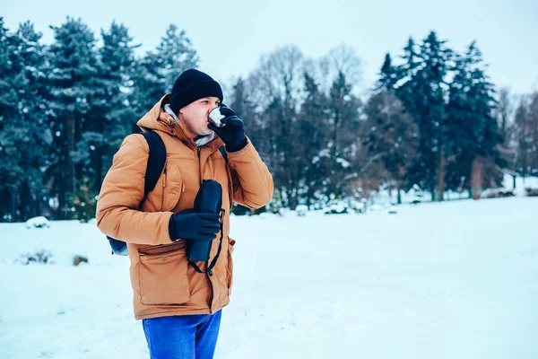 kar üzerinde duran ve bir termos ve bir fincan çay tutan bir sırt çantası ile kırmızı ceketli genç bir adam. Kış orman.