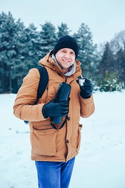 kar üzerinde duran ve bir termos ve bir fincan çay tutan bir sırt çantası ile kırmızı ceketli genç bir adam. Kış orman.