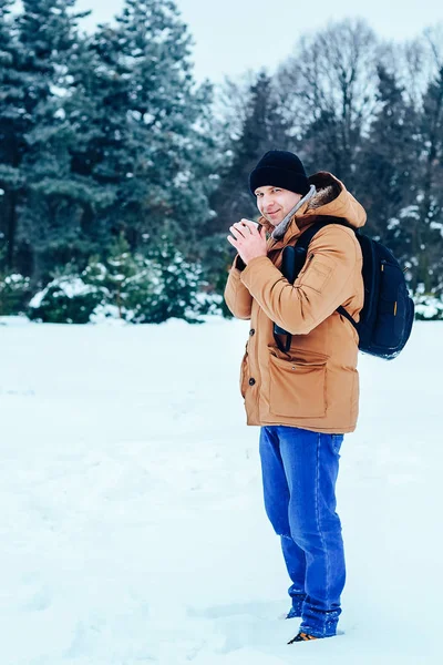kar üzerinde duran ve bir termos ve bir fincan çay tutan bir sırt çantası ile kırmızı ceketli genç bir adam. Kış orman.