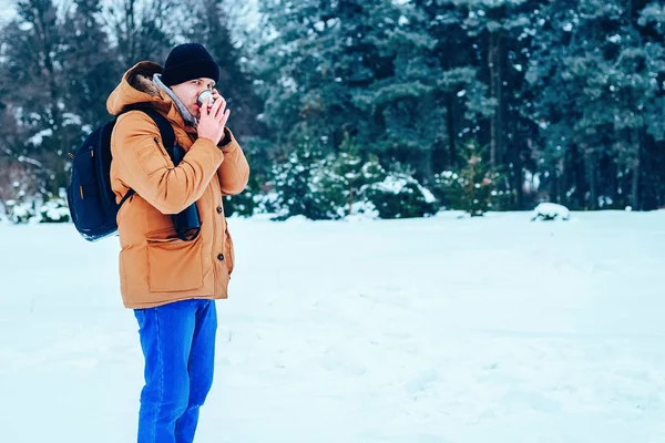 kar üzerinde duran ve bir termos ve bir fincan çay tutan bir sırt çantası ile kırmızı ceketli genç bir adam. Kış orman.