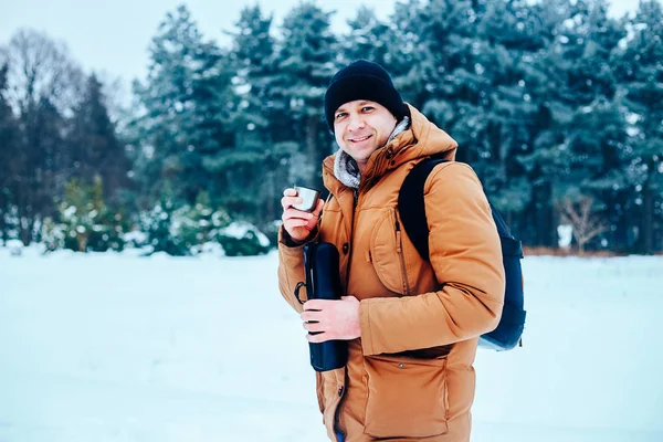 kar üzerinde duran ve bir termos ve bir fincan çay tutan bir sırt çantası ile kırmızı ceketli genç bir adam. Kış orman.