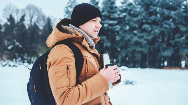 kar üzerinde duran ve bir termos ve bir fincan çay tutan bir sırt çantası ile kırmızı ceketli genç bir adam. Kış orman.