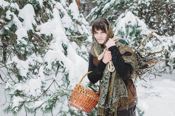 Uma jovem mulher em roupas velhas está de pé com uma cesta e mato na neve na floresta de inverno . — Fotografia de Stock