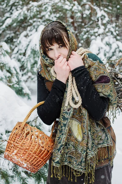 Uma jovem mulher em roupas velhas está de pé com uma cesta e mato na neve na floresta de inverno . — Fotografia de Stock