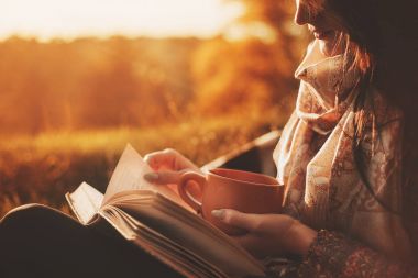 bir kadın bir ağaçta bir sonbahar Parkı yakınındaki oturur ve bir kitap ve bir fincan sıcak bir içecek ile onun elinde tutar. Kız bir kitap okuma