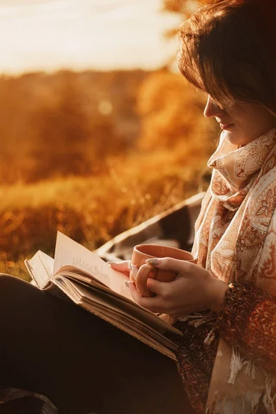 Kobieta siedzi w pobliżu drzewa w parku jesień i posiada książkę i kubek z kubkiem gorącego napoju w jej ręce. Dziewczyna z książką — Zdjęcie stockowe
