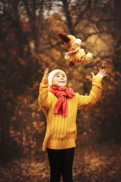 Holčička ve žlutém svetru a červeným šátkem stojí na pozadí podzimního lesa a hází hračky pes — Stock fotografie