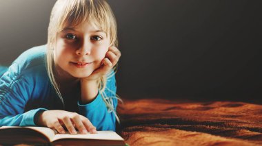 küçük sarışın kız mavi bluz içinde karanlık bir odada yatıyor ve bir yeşil kitap okur