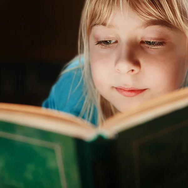 สาวผมบลอนด์น้อยในเสื้อคลุมสีฟ้า นอนอยู่ในห้องมืดและอ่านหนังสือสีเขียว — ภาพถ่ายสต็อก