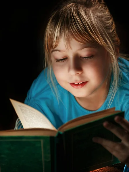 Маленька блондинка в блакитній блузці лежить у темній кімнаті і читає зелену книгу — стокове фото