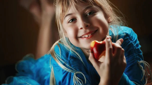 A little blonde girl in a blue blouse lies in a dark room and eats an apple. The girl is holding a biting apple — Stock Photo, Image