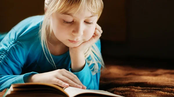 Blond dziewczynka w niebieska bluzka leży w ciemnym pokoju i czyta książkę zielony — Zdjęcie stockowe