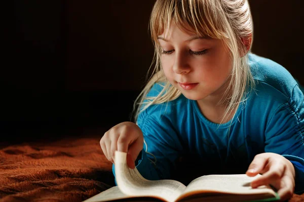 Маленька блондинка в блакитній блузці лежить у темній кімнаті і читає зелену книгу — стокове фото