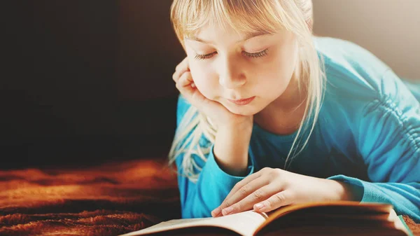 Een blond meisje in een blauwe blouse ligt in een donkere kamer en leest een groene boek — Stockfoto