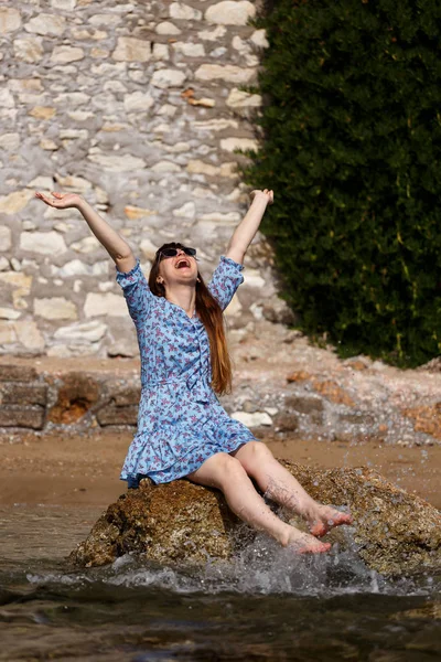 Retrato de mulher junto ao mar. Uma menina com cabelo escuro, em óculos de sol e uma placa de circuito azul senta-se em uma pedra no mar — Fotografia de Stock