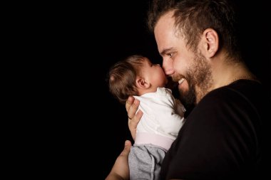Siyah tişörtlü yakışıklı kaslı adamın stüdyo portresi. Siyah arka planlı şirin bir bebeğin ellerinde. Aile, aşk ve mutluluk kavramı.