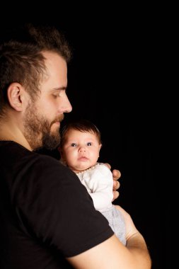 Siyah tişörtlü yakışıklı kaslı adamın stüdyo portresi. Siyah arka planlı şirin bir bebeğin ellerinde. Aile, aşk ve mutluluk kavramı.