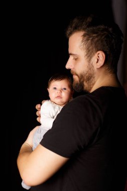 Siyah tişörtlü yakışıklı kaslı adamın stüdyo portresi. Siyah arka planlı şirin bir bebeğin ellerinde. Aile, aşk ve mutluluk kavramı.