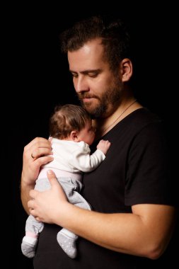 Siyah tişörtlü yakışıklı kaslı adamın stüdyo portresi. Siyah arka planlı şirin bir bebeğin ellerinde. Aile, aşk ve mutluluk kavramı.