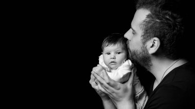 Siyah tişörtlü yakışıklı kaslı adamın stüdyo portresi. Siyah arka planlı şirin bir bebeğin ellerinde. Aile, aşk ve mutluluk kavramı.