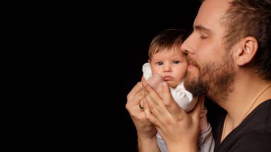 Siyah tişörtlü yakışıklı kaslı adamın stüdyo portresi. Siyah arka planlı şirin bir bebeğin ellerinde. Aile, aşk ve mutluluk kavramı.