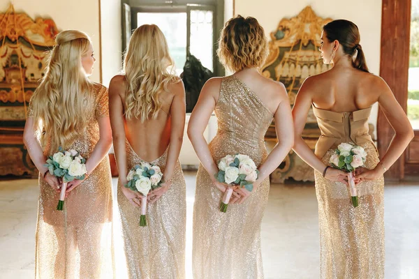 four bridesmaids in golden dresses are standing with their backs and holding bouquets in the hall.
