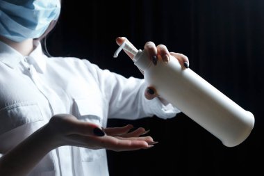 Siyah arka planda bir şişe antiseptik tutan kadın ellerini kapatın. Tıbbi maskeli ve beyaz bluzlu bir kadın antiseptik kullanıyor.