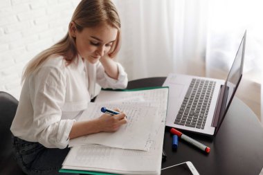 Beyaz bluzlu genç sarışın kız odada çalışıyor. Belgeleri ve dizüstü bilgisayarı var. Evde çalış. Kadın, elinde belgelerle masada otururken kameraya bak..