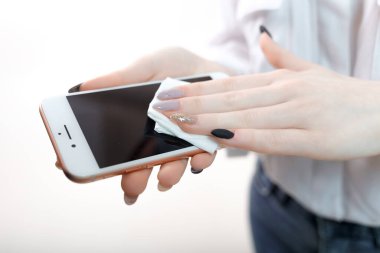 Beyaz bluzlu ve tıbbi maskeli genç sarışın bir kız beyaz arka planda antiseptik peçeteyle akıllı telefonu siler. İnsan elleri telefonu spritik antiseptik mendille siler. Akıllı telefon dezenfeksiyonu. kadın elleri