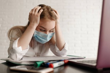 Beyaz bluzlu ve tıbbi maskeli genç sarışın kız odada evrak ve dizüstü bilgisayarla çalışıyor. Evde çalış. Bir kadın masaya oturur ve ellerini başının arkasında tutar. Çalışmaktan ve kendini soyutlamaktan yoruldum.