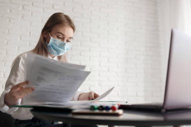 Beyaz bluzlu ve tıbbi maskeli genç sarışın kız odada evrak ve dizüstü bilgisayarla çalışıyor. Evde çalış. Bir kadın masaya oturur ve elinde bir kağıt tutar.