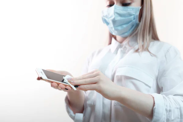 young blonde girl in a white blouse and in a medical mask wipes a smartphone with an antiseptic napkin on a white background.human hands wipe the phone with a spritic antiseptic wipe. smartphone disinfection. female hands