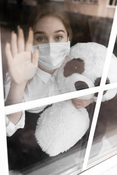 a young blonde girl in a white blouse and a medical mask sits outside the window and looks outside. Quarantine and home isolation from coronavirus.a young blonde girl in a white blouse and a medical mask sits outside the window and looks outside. Gir