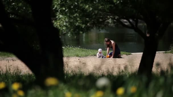 幸せな母と湖のビーチで遊ぶ女の子の赤ん坊 タンポポで草を撮影します 女の赤ちゃん生後 水の背景 — ストック動画